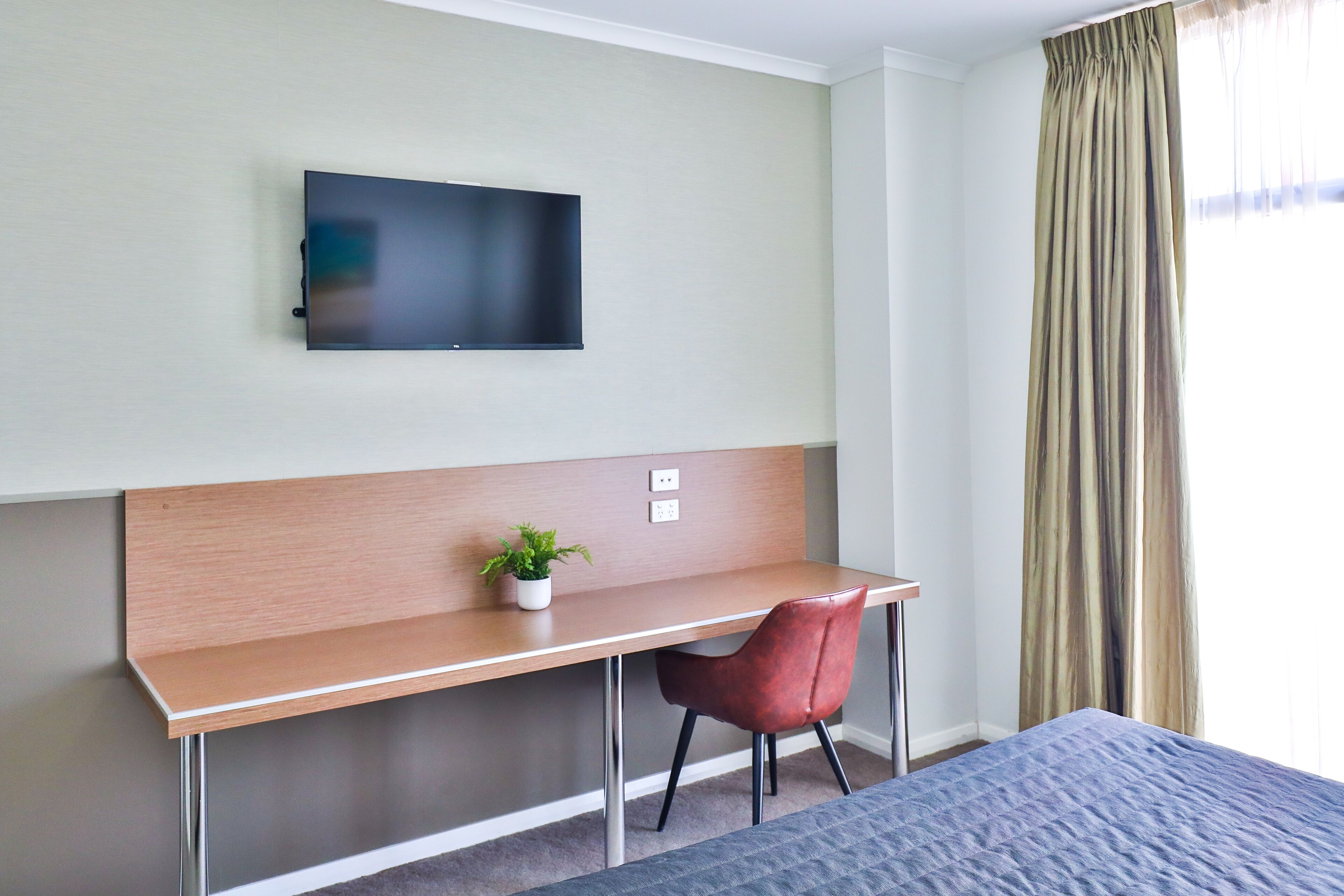 A hotel room with a bed, a desk with a chair, a television on the wall, and a window with curtains.