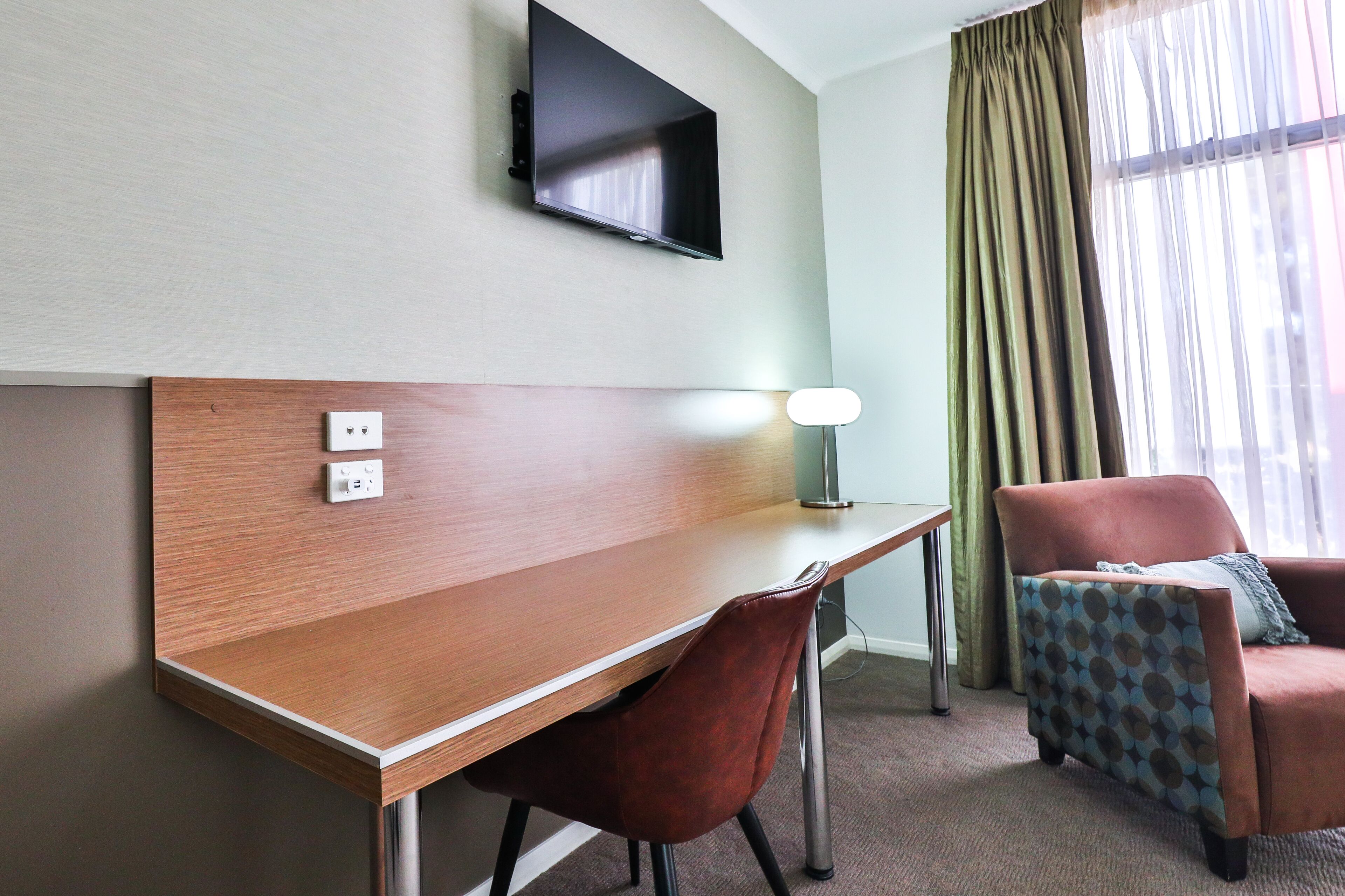 Ett hotellrum med ett skrivbord i trä, en röd stol, en tv som är monterad på väggen och en fåtölj med mönster.