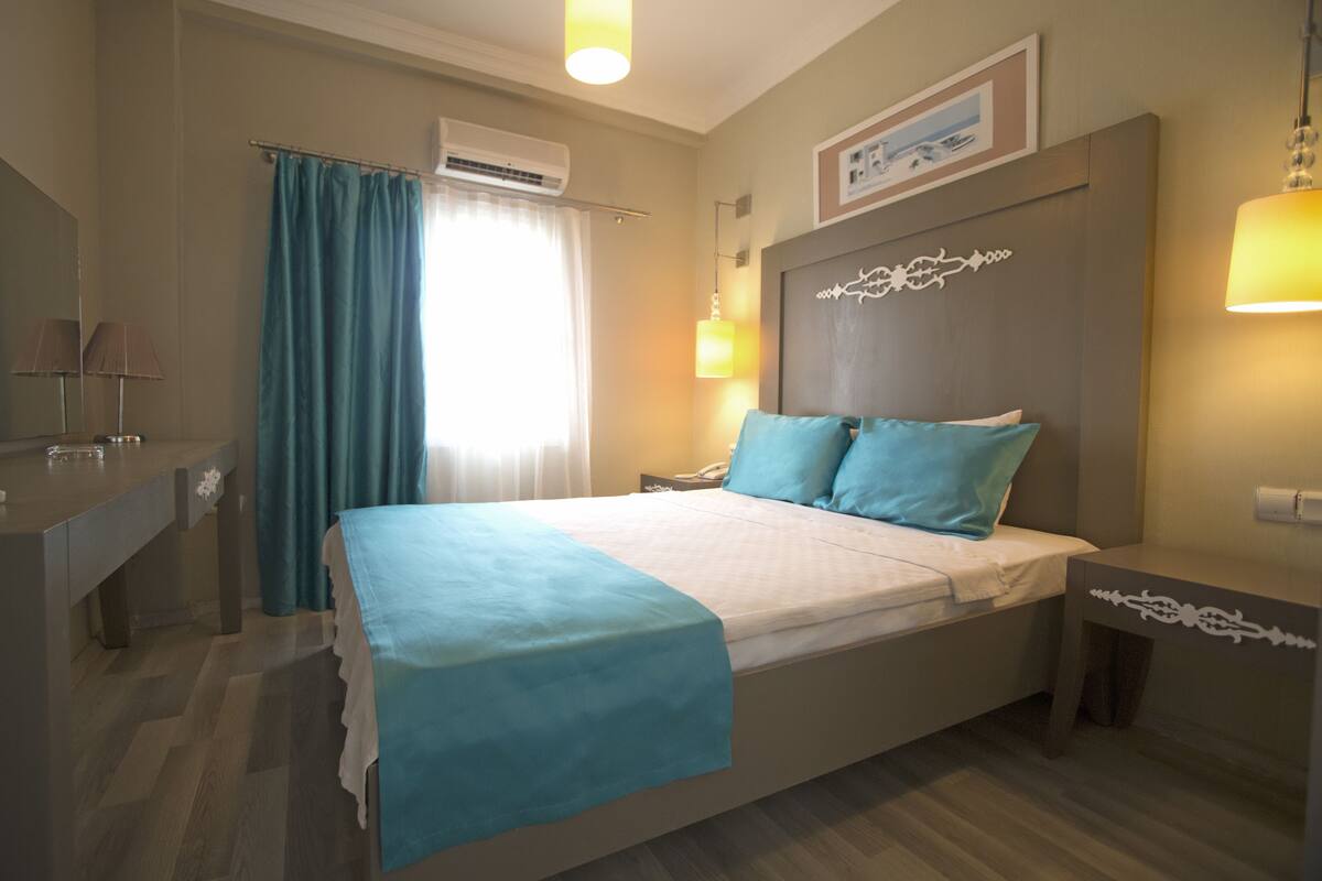 Quarto de hotel com cama, mesas de cabeceira, escrivaninha e janela com cortinas.