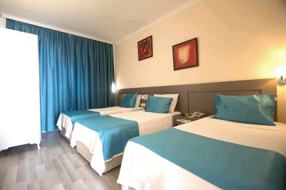 Quarto de hotel com três camas, cada uma com colcha azul-turquesa e lençóis brancos. Há duas imagens emolduradas na parede, uma mesa de cabeceira com telefone e uma janela com cortinas azuis.