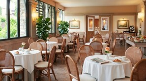 A dining area with round tables, wicker chairs, and a view of greenery through large windows.