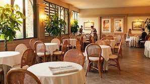 Un restaurante con mesas redondas, sillas de mimbre, ventanas amplias y plantas de interior.