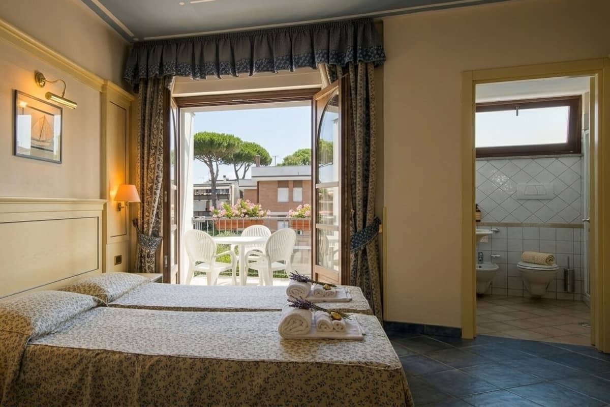 A hotel room with a bed, bedside lamps, a telephone, a balcony with chairs, and a view of trees and buildings.