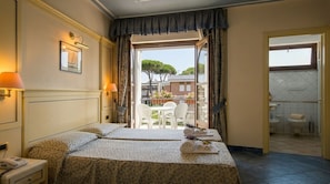 A hotel room with a bed, bedside lamps, a telephone, a balcony with chairs, and a view of trees and buildings.