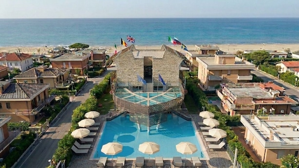 Un hotel con piscina central, rodeado de sillas de descanso y sombrillas, con banderas en la parte superior y vista a la playa.
