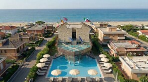 Un hotel con piscina central, rodeado de sillas de descanso y sombrillas, con banderas en la parte superior y vista a la playa.