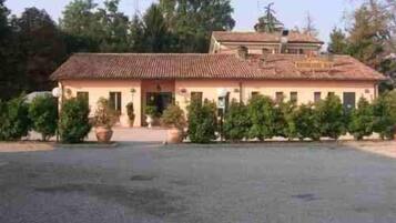Un edificio a due piani con tetto in tegole, circondato da vegetazione e un'area pavimentata antistante.