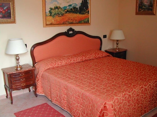 Un dormitorio con una cama, dos mesitas de noche con lámparas, un tapete rojo y dos cuadros enmarcados en la pared.