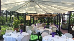 Un área de comedor al aire libre con mesas dispuestas para una comida, rodeada de follaje y una estructura cubierta.