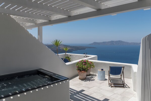 A balcony with a hot tub, potted plants, and a chair overlooking a scenic view of the sea and distant land.