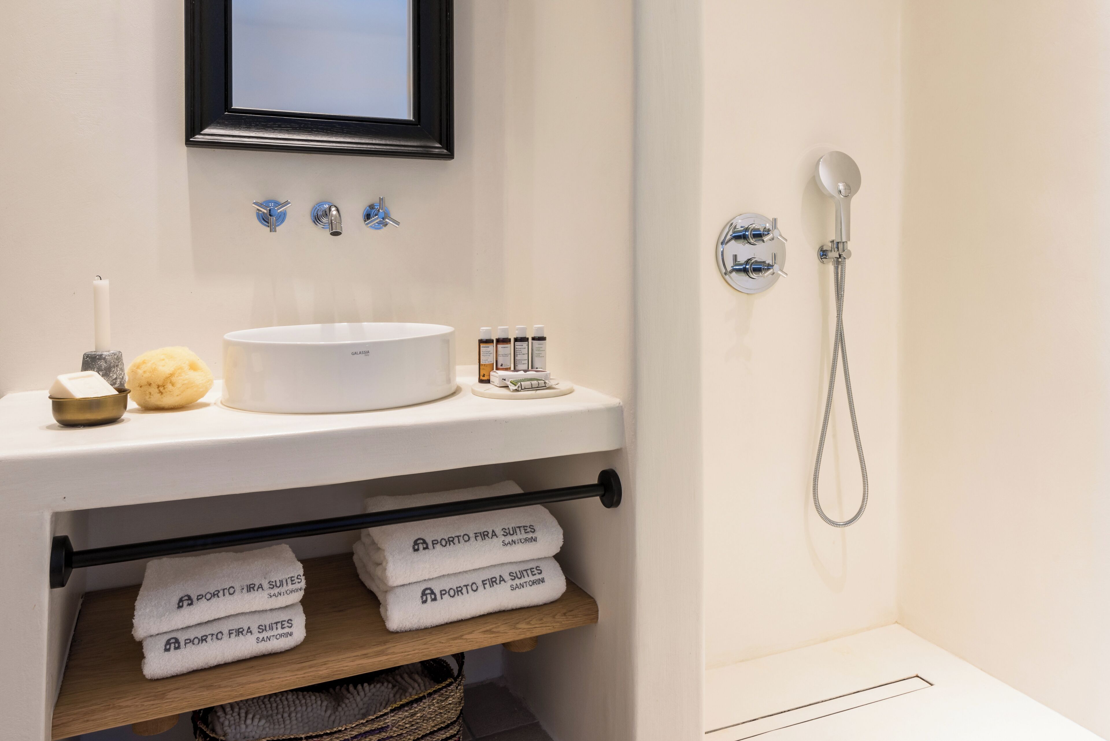 A bathroom with a white sink, a mirror, a framed picture, a shower, and neatly folded towels on a shelf.