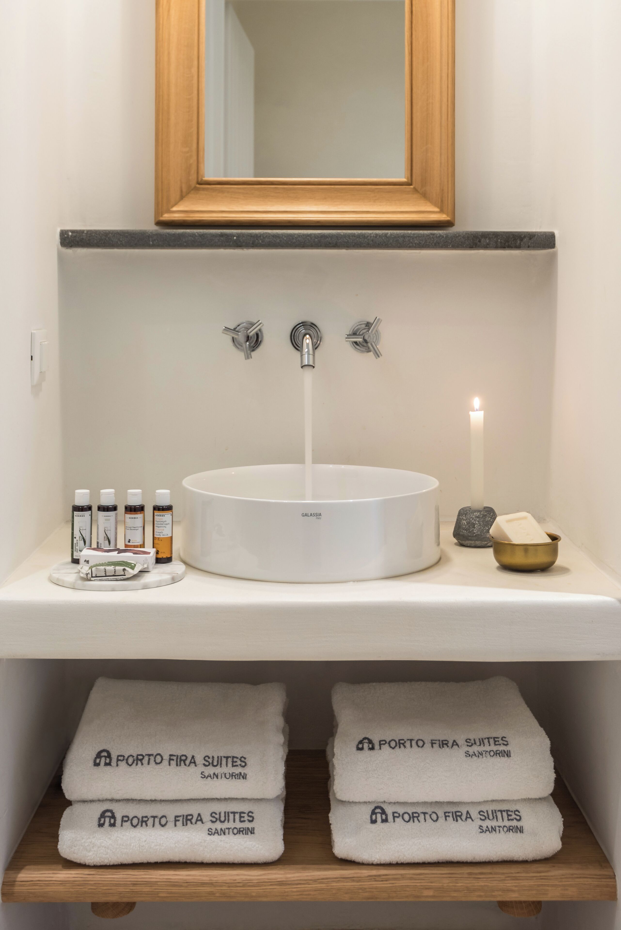 A bathroom with a round white sink, two faucets, a mirror, and a lit candle. There are toiletries on the counter and towels on the shelf below.