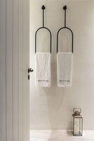 Dos toallas blancas colgadas en ganchos negros, con la inscripción "PORTO FIRA SUITES" y un pequeño farol de cristal en el suelo.