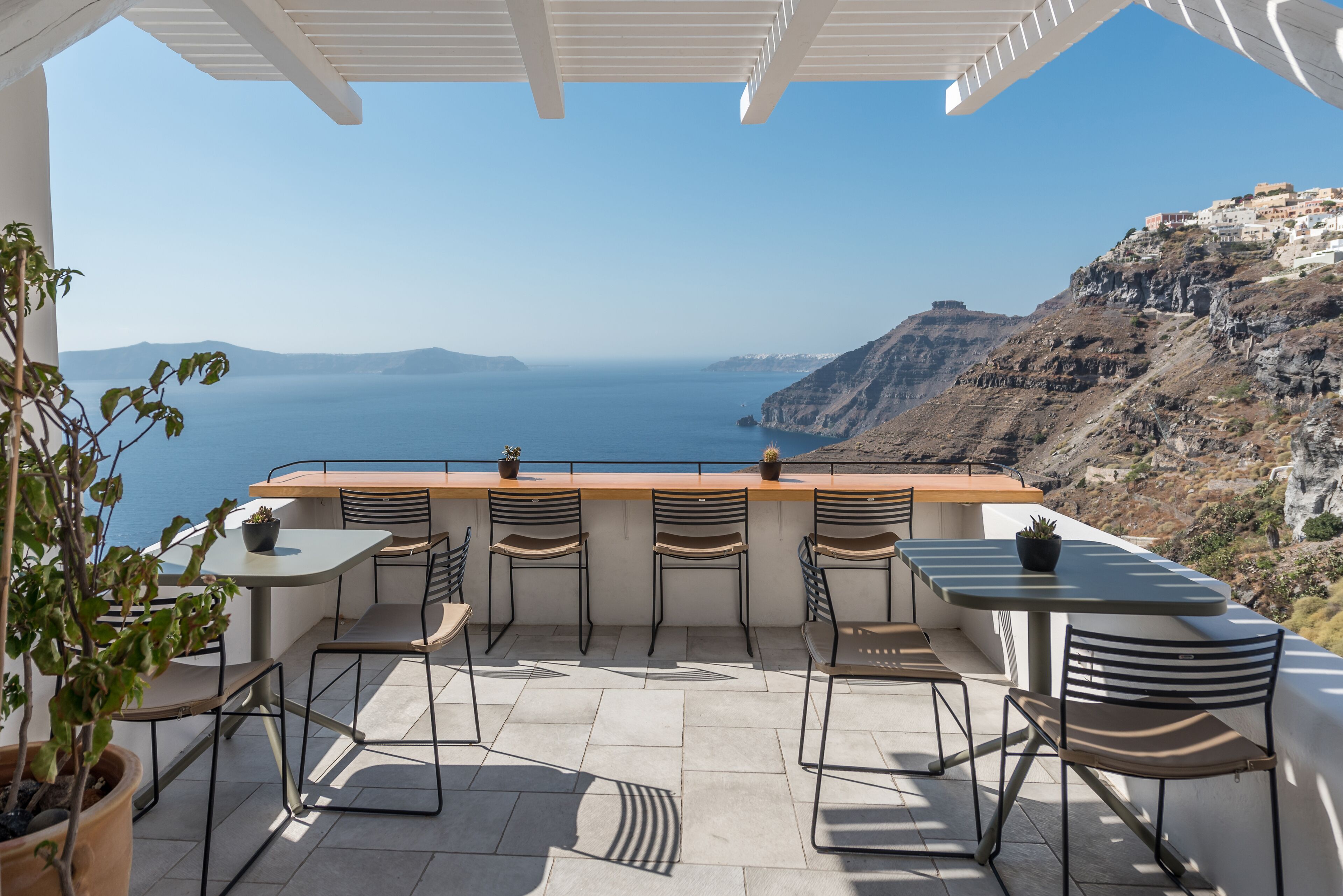An outdoor terrace with a view of the sea and mountains, featuring a wooden table and chairs, potted plants, and a pergola.