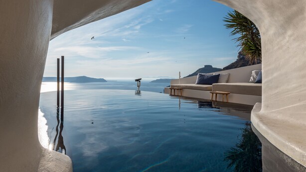 A infinity pool with a clear view of the sea and distant mountains, a white infinity wall, and a seating area with cushions. - Porto Fira Suites (Santorini)