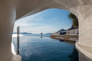 A infinity pool with a clear view of the sea and distant mountains, a white infinity wall, and a seating area with cushions.