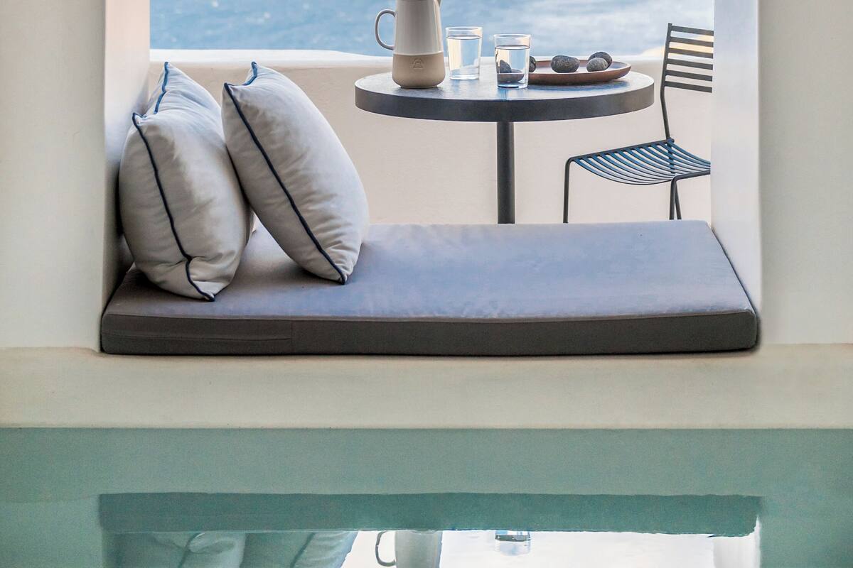 A balcony with a view of the sea, a table with a mug, glasses, and a plate of fruit, and two pillows.