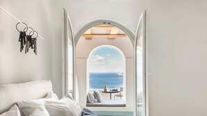 A bedroom with a view of the sea through an arched window, a bed with pillows, and a dreamcatcher on the wall. - Porto Fira Suites (Santorini)