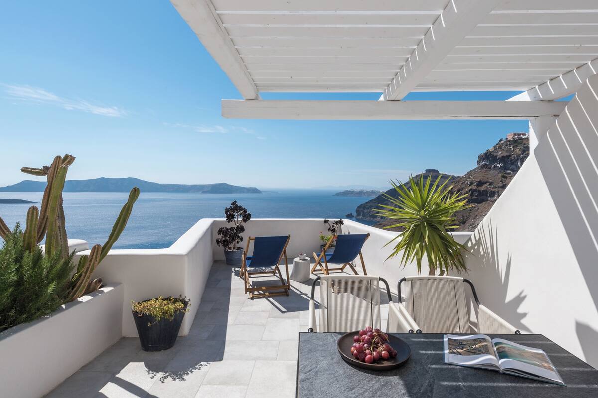 A terrace with a view of the sea, featuring a table with a book and grapes, two blue chairs, and potted plants.