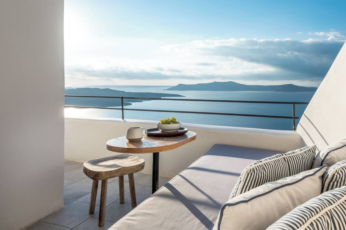 A balcony with a view of the sea and mountains, a small table with a bowl of fruit, and a cushioned seating area.
