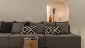 A modern living room with a grey sectional sofa, patterned throw pillows, a wooden coffee table, and a wall-mounted floor lamp. - Porto Fira Suites (Santorini)