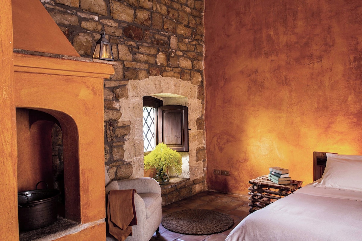 A rustic room with a stone wall, a fireplace, a bed, a small window with a view, and a wooden side table with books.