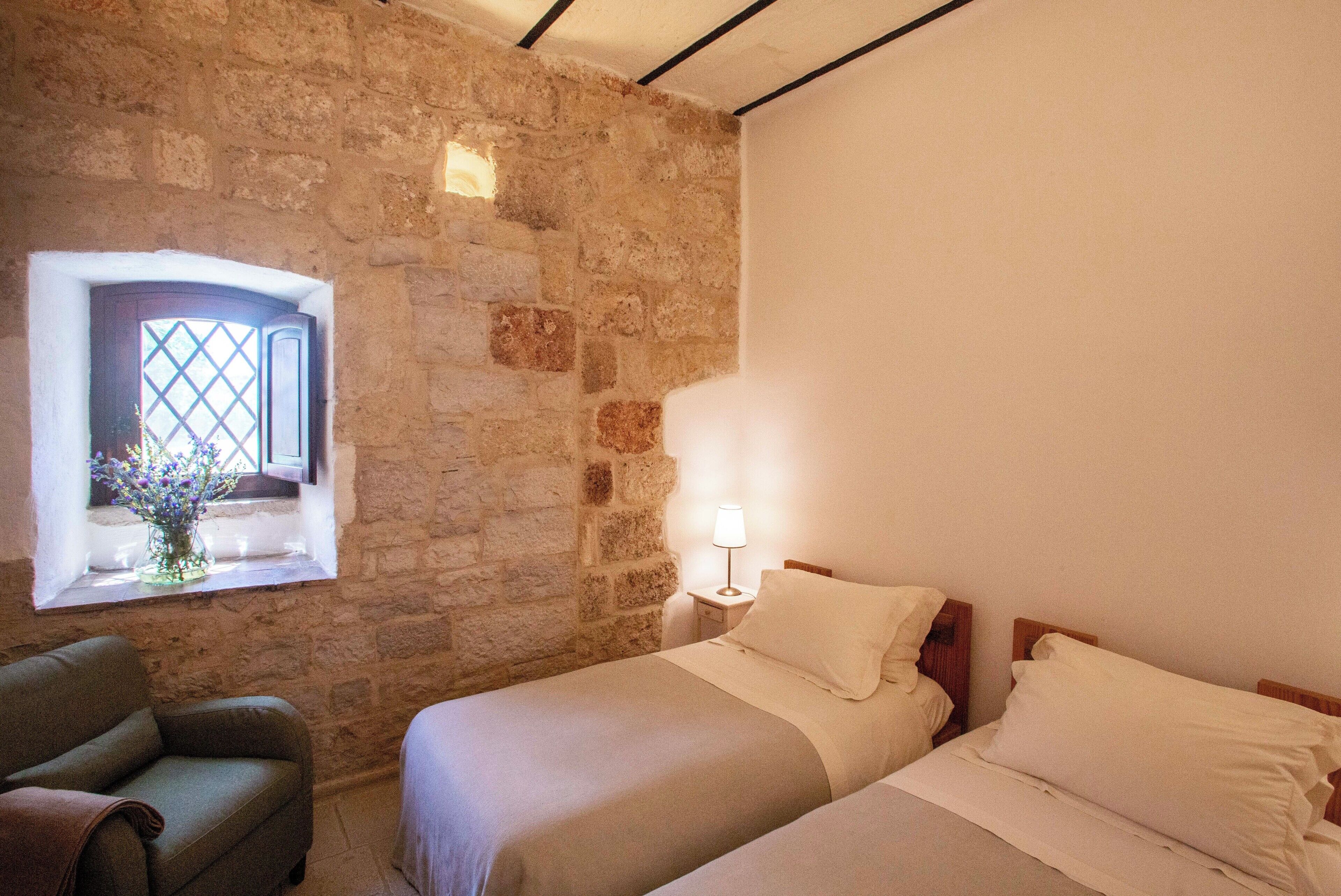 Quarto com parede de pedra, janela com vista, cama, mesa de cabeceira com abajur e uma cadeira.