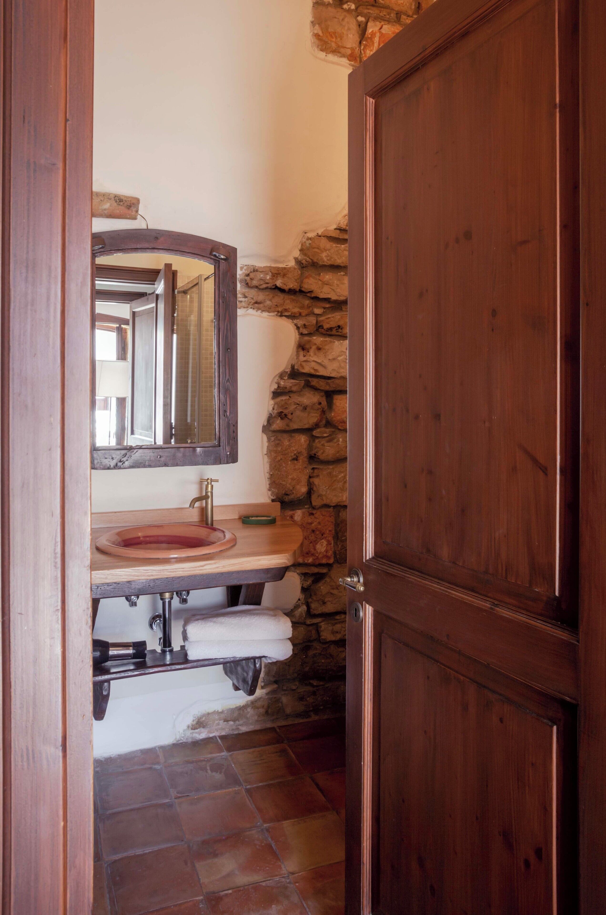 Un baño con pared de piedra, puerta de madera, lavabo con espejo y perchero para toallas.