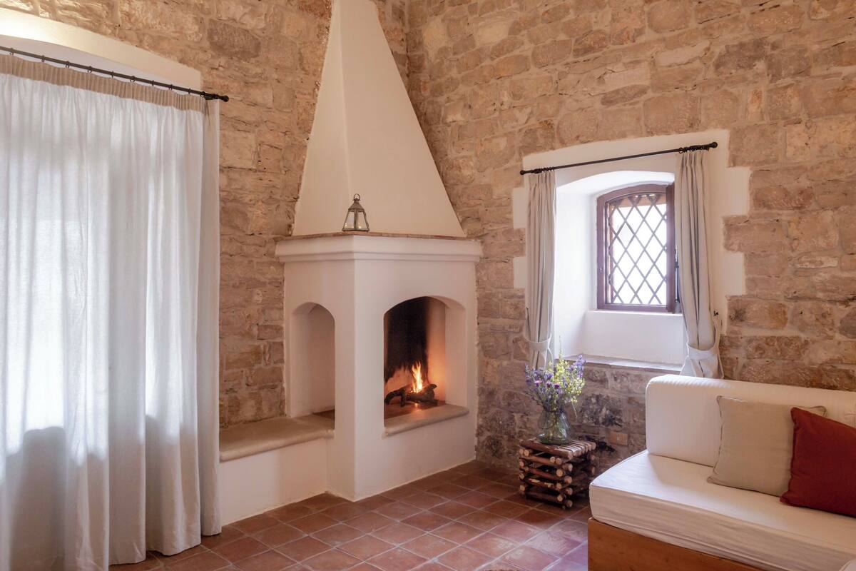 A cozy room with a fireplace, stone walls, a white sofa, and a window with curtains.
