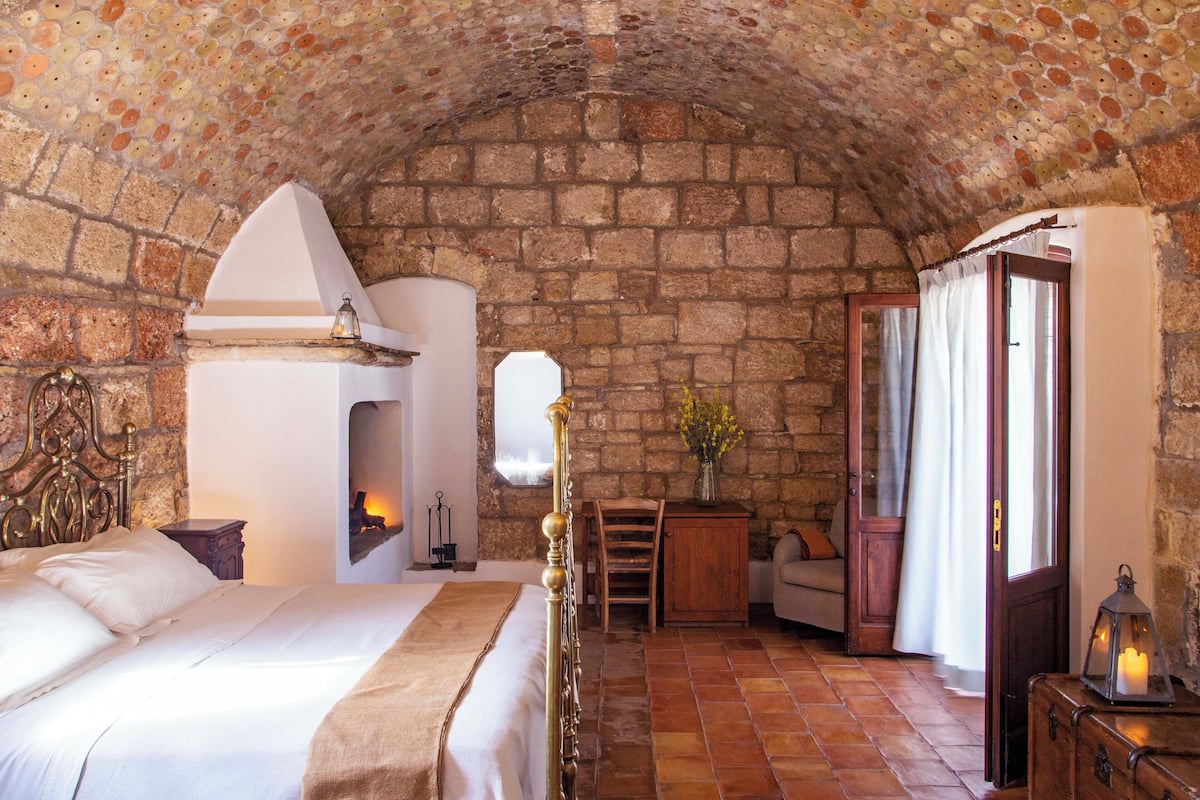 A cozy room with a stone wall, a fireplace, a bed, a wooden chair, and a small table with a vase of flowers.