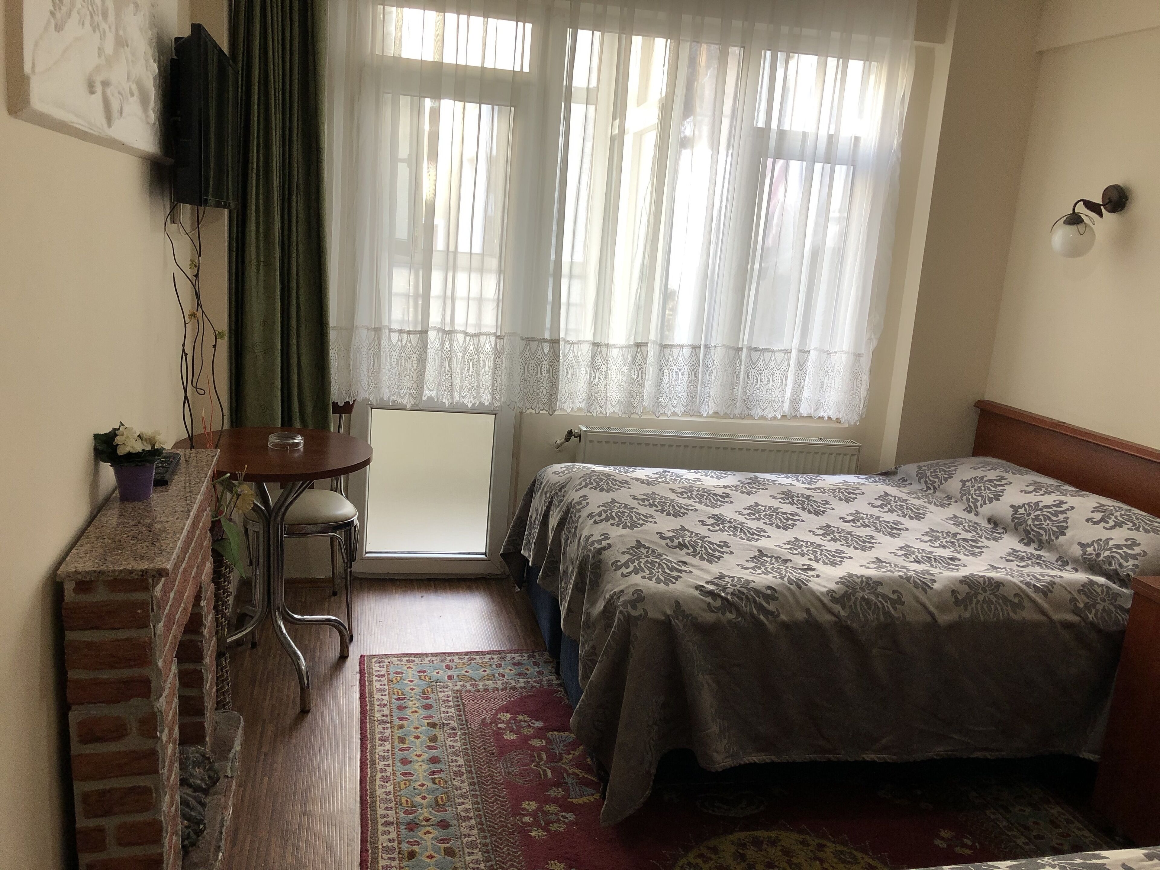 A bedroom with a bed, a small table, a chair, a radiator, and a window with curtains.