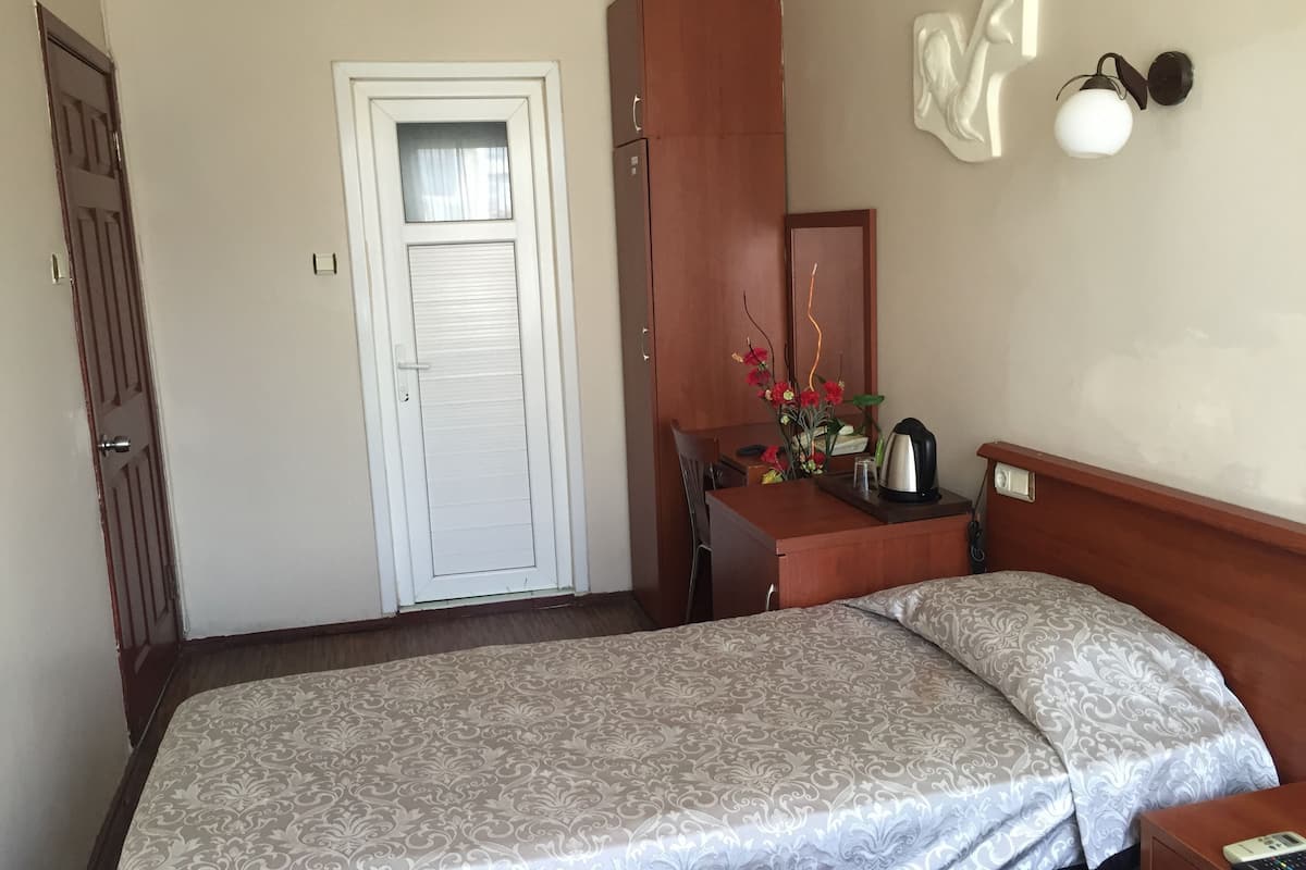 A bedroom with a bed, a wooden desk, a chair, a mirror, a wall-mounted light fixture, and a door.