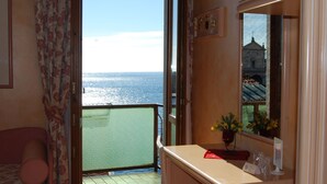 Habitación con balcón con vista al mar, cama, una mesita con un florero y un espejo que refleja un edificio.