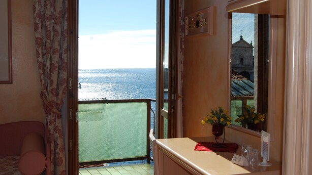 A room with a balcony overlooking the sea, a bed, a small table with a vase of flowers, and a mirror reflecting a building. - Garda Sol Apart-hotel Beauty & SPA (Toscolano Maderno)