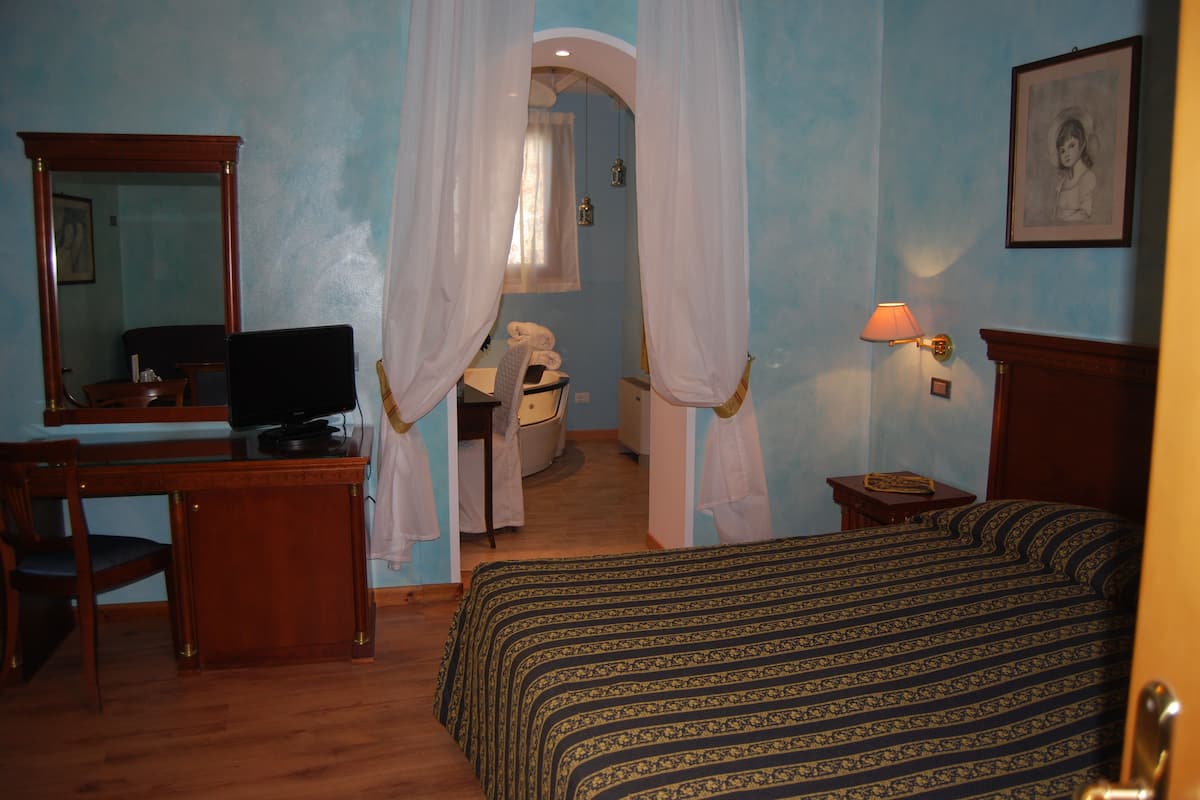 A bedroom with a bed, a desk, a chair, a television, and a bathroom visible through a doorway.