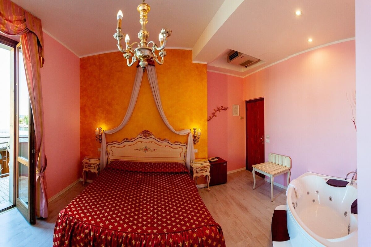 A bedroom with a four-poster bed, a chandelier, a bathtub, and a balcony with curtains.
