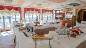 A buffet setup with various food items and utensils, a chandelier, and large windows with curtains. - Garda Sol Apart-hotel Beauty & SPA (Toscolano Maderno)