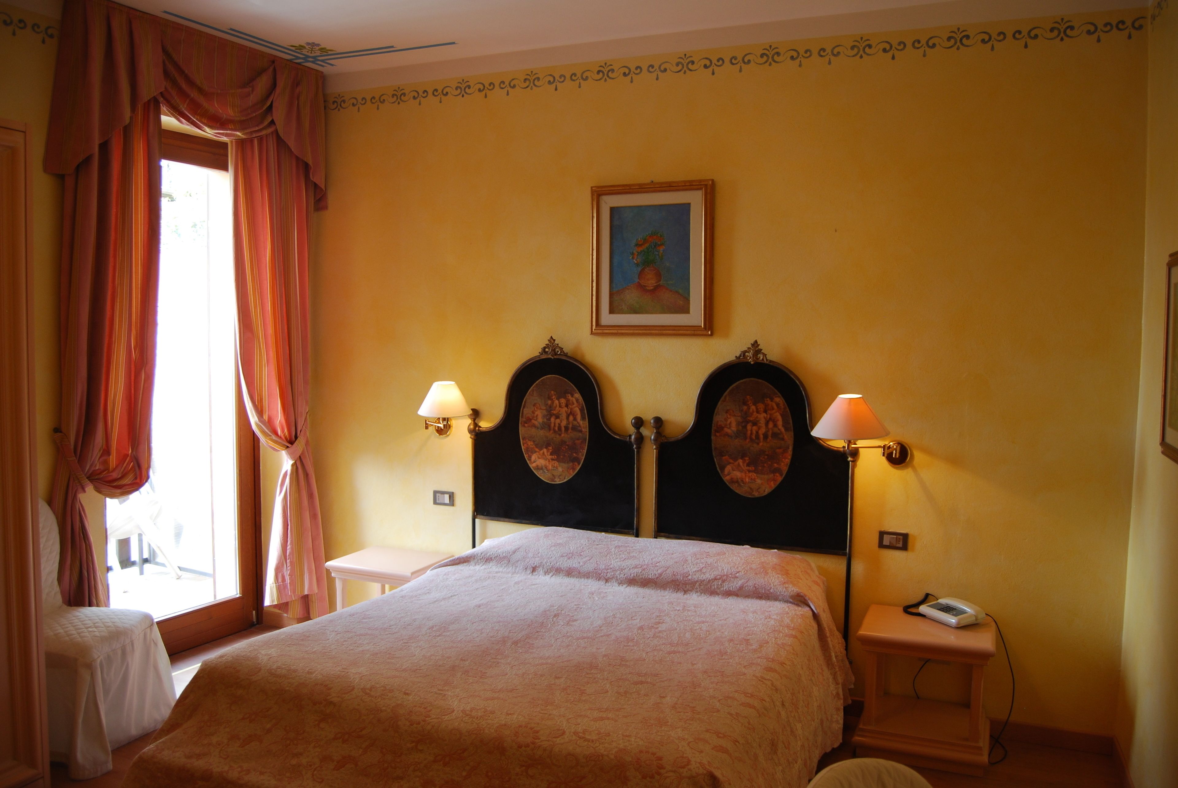 A hotel room with a bed, two bedside lamps, a nightstand, a painting on the wall, and a window with curtains.