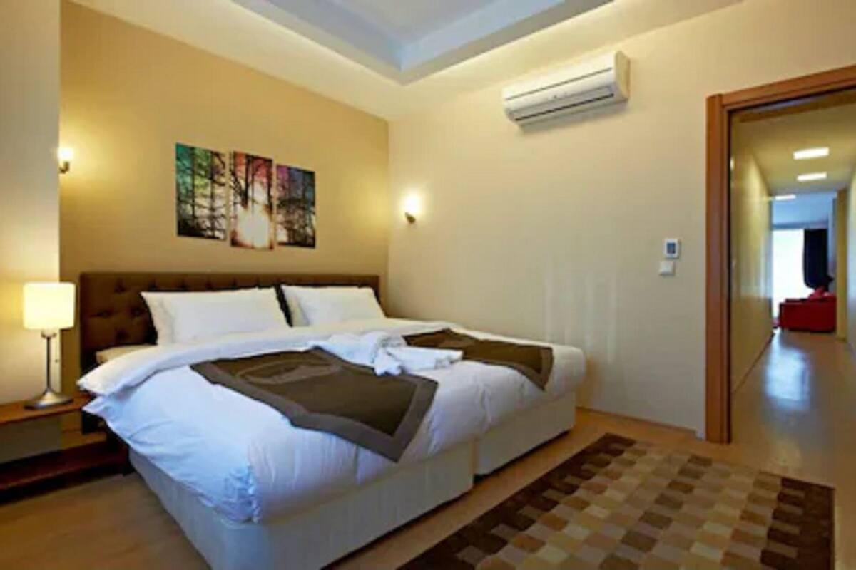 A hotel room with a large bed, a nightstand, a lamp, an air conditioner, and a painting on the wall.