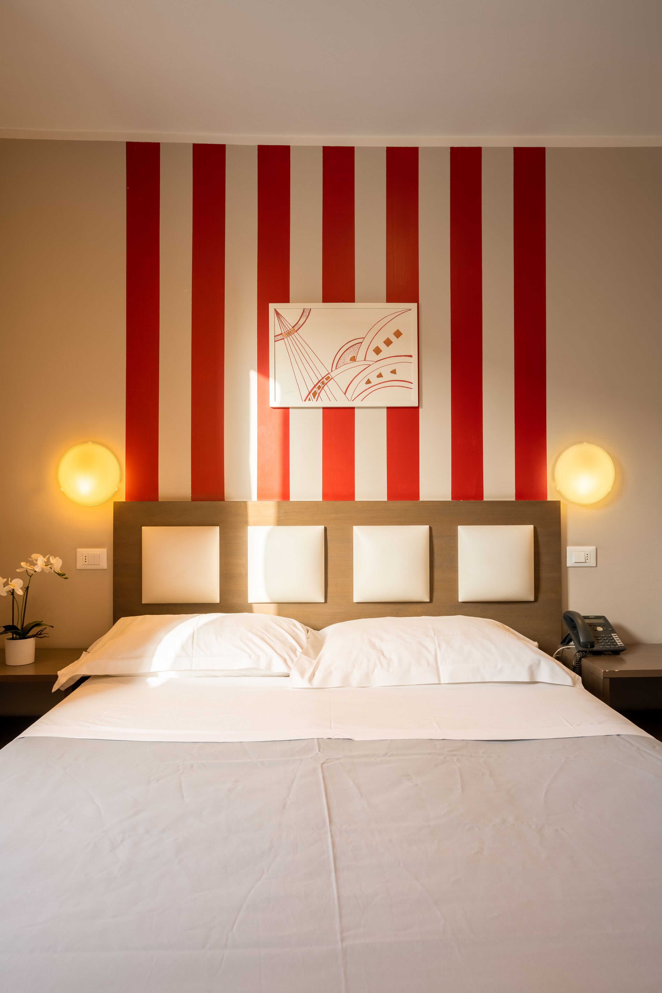 Habitación de hotel con cama, pared a rayas, lámpara de pared y una mesita con un teléfono.