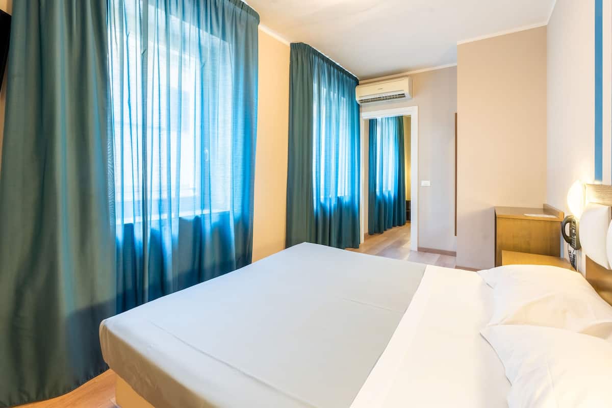 A hotel room with a large bed, a bedside table, a lamp, and a view of the hallway.