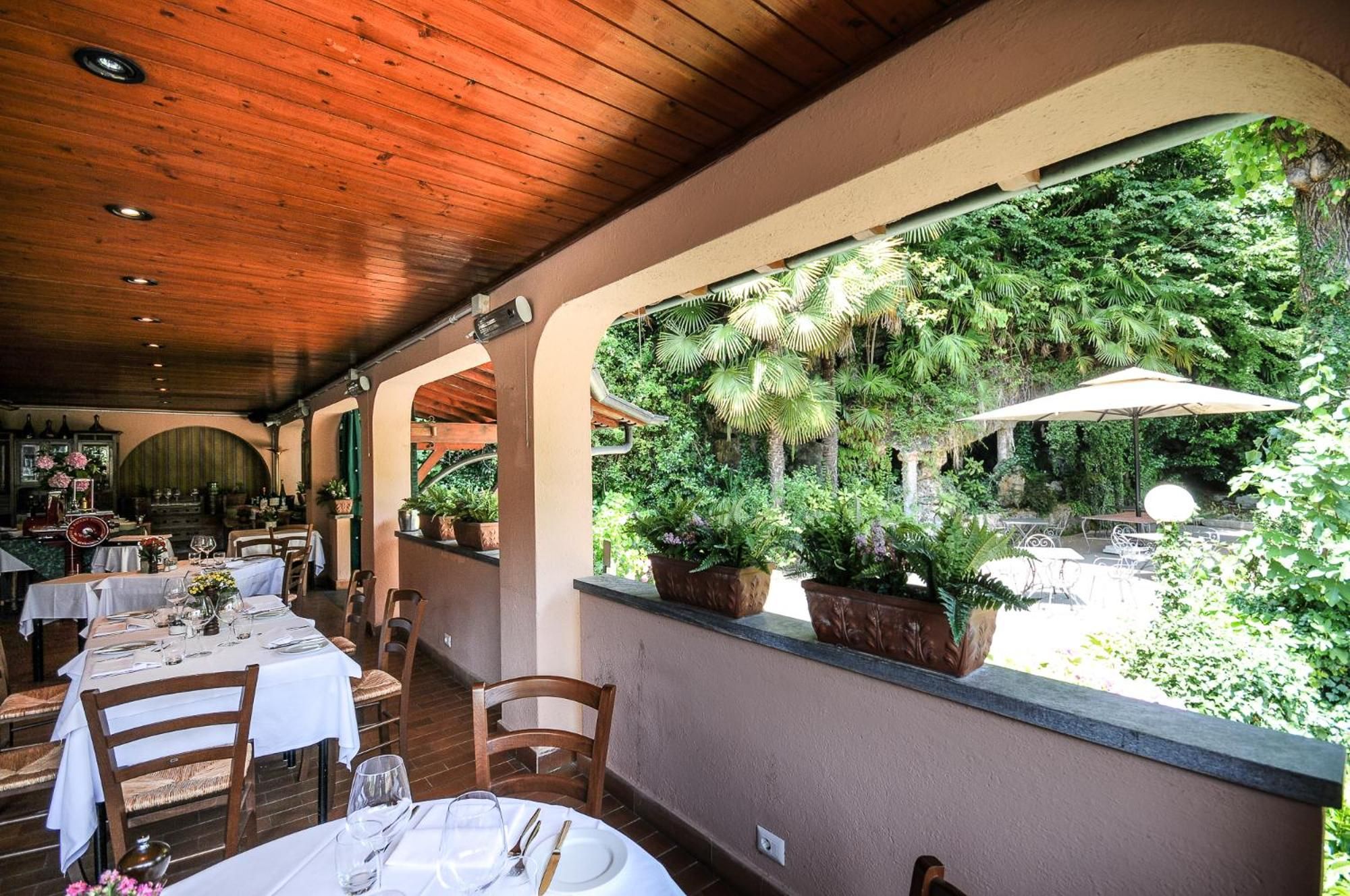 Um espaço de refeições interno com mesas para dois, teto de madeira e vista para um jardim exuberante através de grandes janelas.
