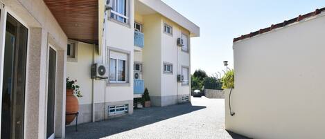A narrow street leading to a building with multiple balconies, a car parked in front, and a clear sky.