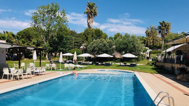 Una piscina con sillones de playa y sombrillas, rodeada de vegetación y un cielo despejado.