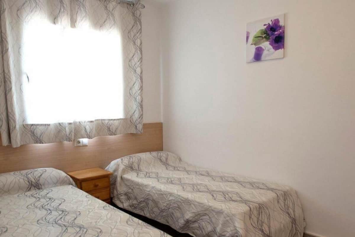 A room with two beds, a wooden headboard, a small wooden nightstand, and a window with sheer curtains.