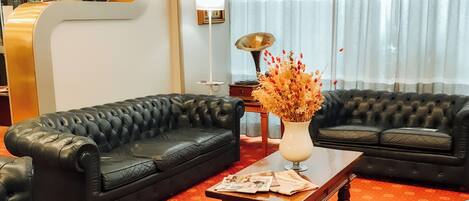 A living room with two black leather sofas, a wooden coffee table with newspapers, a lamp, and a vase with dried flowers.