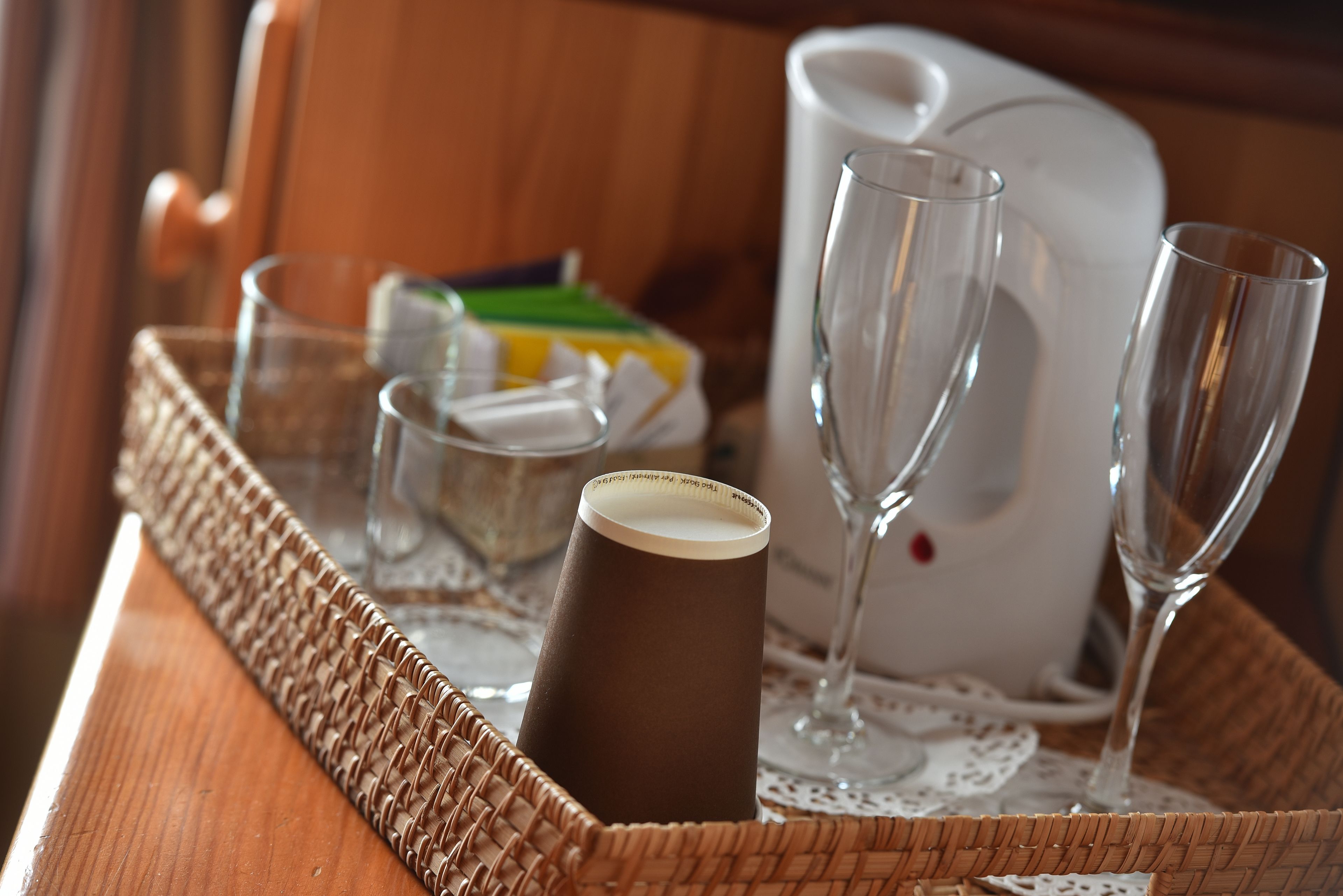 Una bandeja de mimbre con una taza de café, dos copas de vino y una jarra blanca sobre una mesa de madera.