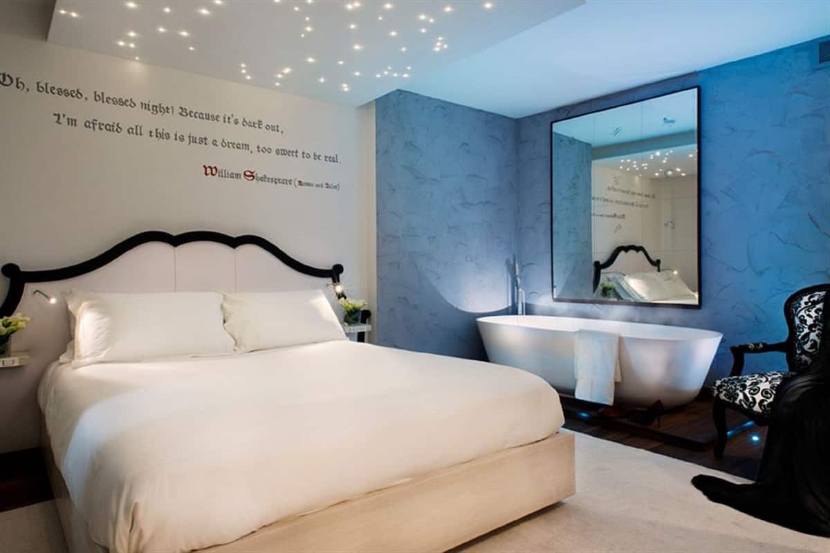 A bedroom with a large bed, a white bedside table with a vase, a modern bathroom with a bathtub and a large mirror, and a quote on the wall.