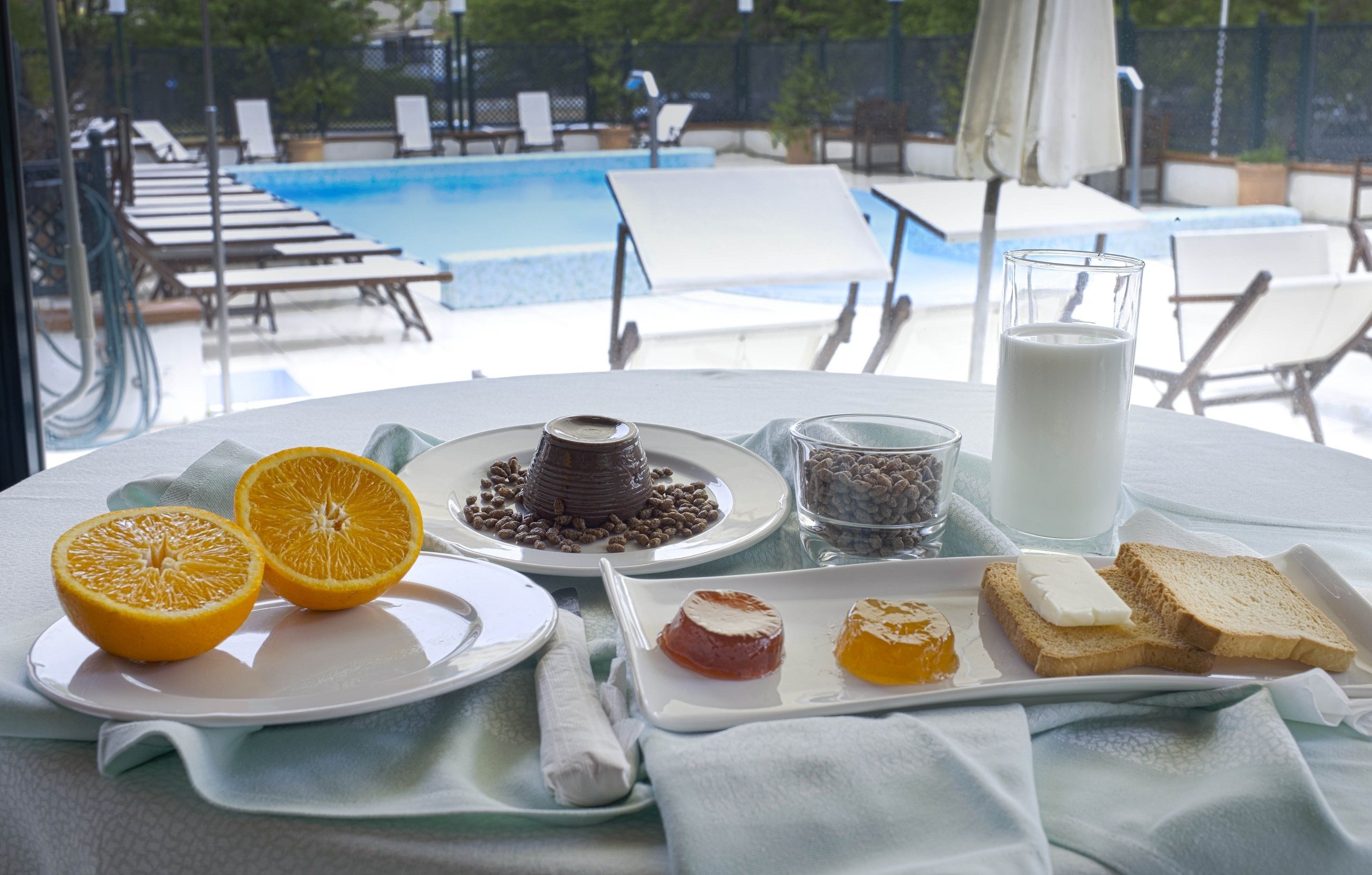 Desayuno con rodajas de naranja, pastel de chocolate, leche y tostadas sobre una mesa con vista a la piscina.