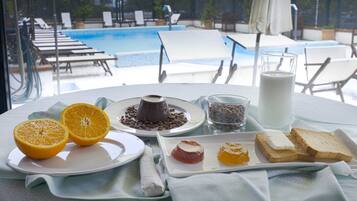 Desayuno con rodajas de naranja, pastel de chocolate, leche y tostadas sobre una mesa con vista a la piscina.